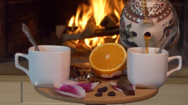 茶话会在壁炉的后面 一个女人把茶壶里的茶倒入小桌子上的杯子里 桌上放着花瓣 巧克力 然后用茶匙搅拌 — 图库视频影像