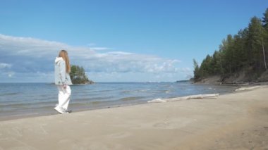 Ağır çekim. Beyaz elbiseli genç bir kadın kumlu sahil boyunca yürüyor. Ob Nehri, Sibirya.
