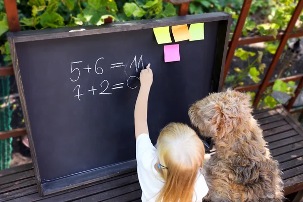 Linda niña con perro escrito respuesta al ejerciciousando tiza en pizarra — Foto de Stock