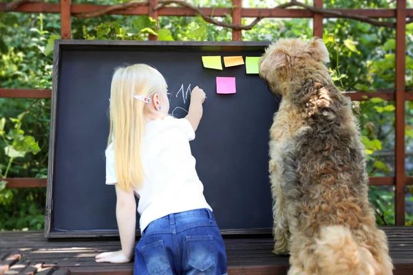 Linda niña con perro escrito respuesta al ejerciciousando tiza en pizarra — Foto de Stock