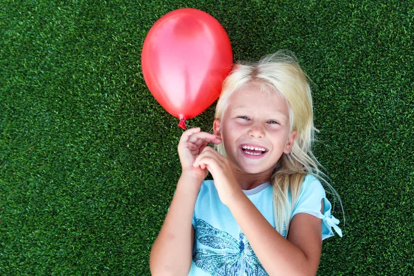 Schönes blondes lächelndes Mädchen, das auf dem Gras liegt und einen roten Ball hält — Stockfoto