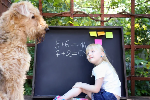 Menina bonito com cão escrevendo resposta para o exercíciousando giz no quadro negro — Fotografia de Stock