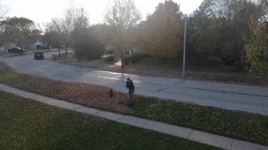 Bir banliyö caddesinde düşen sonbahar yapraklarını toplayan bir adamın hava manzarası. Amerikan banliyö mahallesi. Tek kişilik aile evleri. 4K