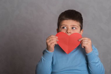Çocuk yüzünü kağıttan kesilmiş büyük bir kırmızı kalbin arkasına sakladı. Çocuk kırmızı kağıttan kesilmiş kalp gösteriyor. Çocuk, kalp şeklinde kırmızı kağıttan bir sevgililer günü kartı tutuyor. Gri arka plandaki bebek aşkın bir sembolünü gösteriyor. Mutlu Çocuk Şovu