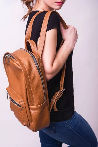 A beautiful, young girl shows a brown, leather backpack in the studio. Leather accessory advertising. Hand luggage. Large, spacious handmade backpack
