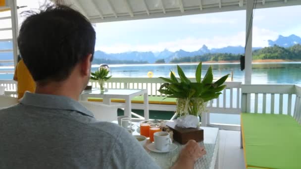 Hombre sentado en el desayuno y beber café en el hermoso lago y las montañas — Vídeo de stock