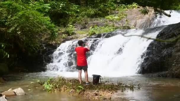 Hombre viajero en la cascada, haciendo fotos o videos en el teléfono móvil en Tailandia — Vídeos de Stock