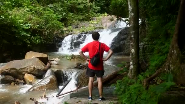 Homem viajante em camiseta vermelha em pé na pedra perto de cachoeira na floresta tropical — Vídeo de Stock