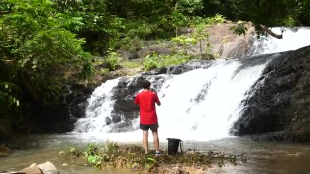 Homem viajante na cachoeira, fazendo foto ou vídeo no celular na Tailândia — Vídeo de Stock