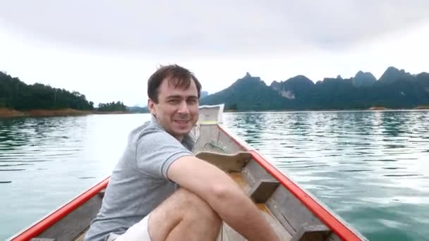 Γελώντας άνθρωπος κάθεται στο ξύλινο σκάφος, μιλώντας και πλέοντας στην πρωινή μπλε λίμνη — Αρχείο Βίντεο
