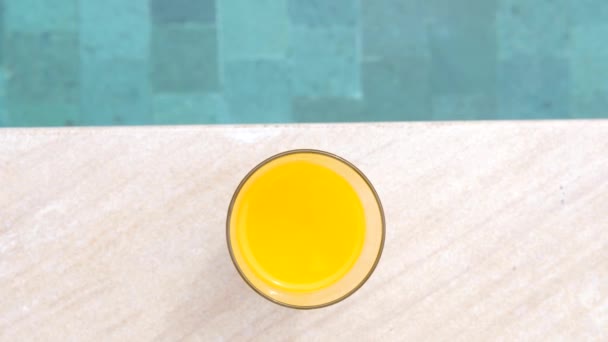 Ovanifrån av glas apelsinjuice stående på kanten av blå pool — Stockvideo