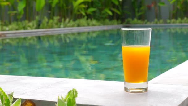 Conceito de férias: Vidro de suco de laranja em pé na borda da piscina azul — Vídeo de Stock