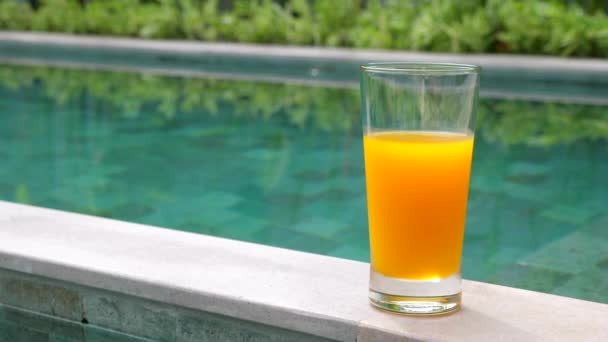 Glas av apelsinjuice stående på kanten av blå pool med tropisk grön — Stockvideo