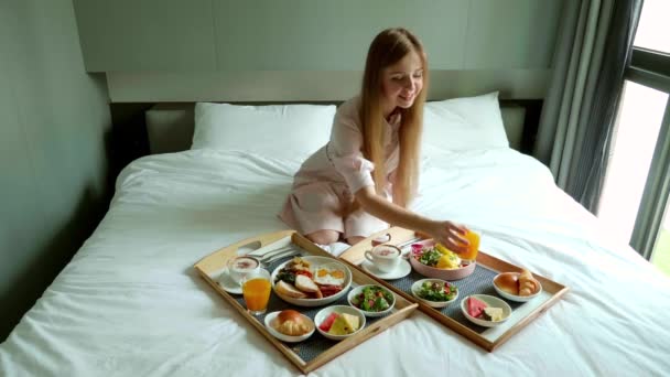 İki tepsi dolusu yiyecek ve portakal suyu ile yatakta oturan gülümseyen kadın. — Stok video