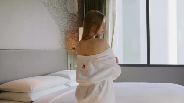 Vue arrière de la femme en peignoir blanc debout près du lit et regardant la fenêtre — Video