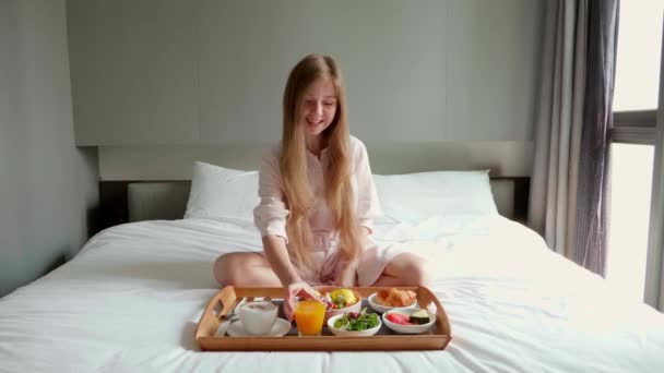 Unga leende vacker kvinna äter frukost i sängen i mysiga hotellrum — Stockvideo