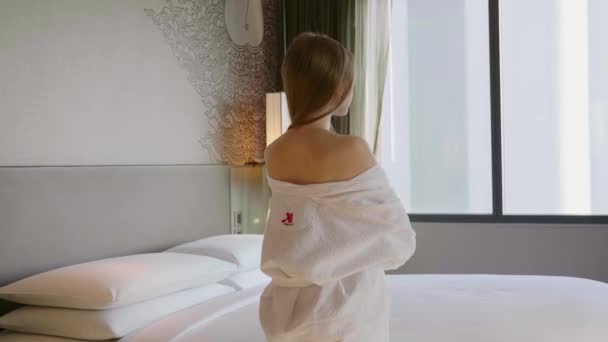 Vue arrière de la femme en peignoir blanc debout près du lit et regardant la fenêtre — Video