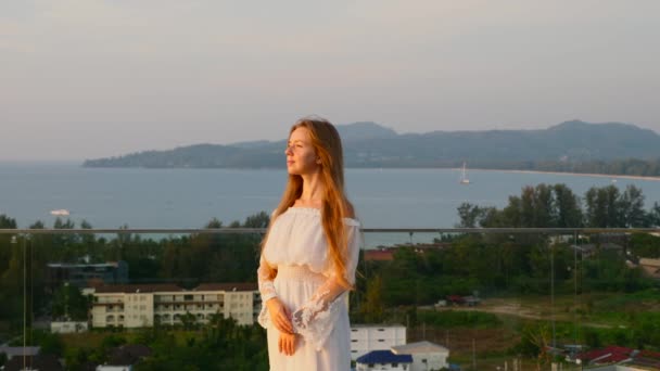 Femme souriante en robe blanche debout sur le toit avec mer, coucher de soleil et montagne — Video