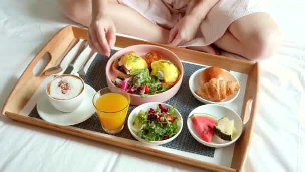 Γυναίκα τρώει πρωινό στο κρεβάτι στο άνετο δωμάτιο του ξενοδοχείου. Πρωινό φαγητό με καπουτσίνο — Αρχείο Βίντεο