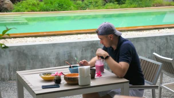 Modern Young Man Sit in Tropical Cafe dekat Green Pool, Read News Feed in Phone — Stok Video