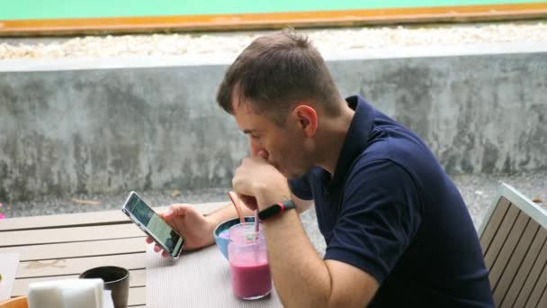 年轻英俊男子在电话里看新闻，喝粉红健康水果滑 — 图库视频影像