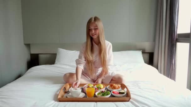 Joven sonriente hermosa mujer comiendo desayuno en la cama en acogedora habitación de hotel — Vídeos de Stock