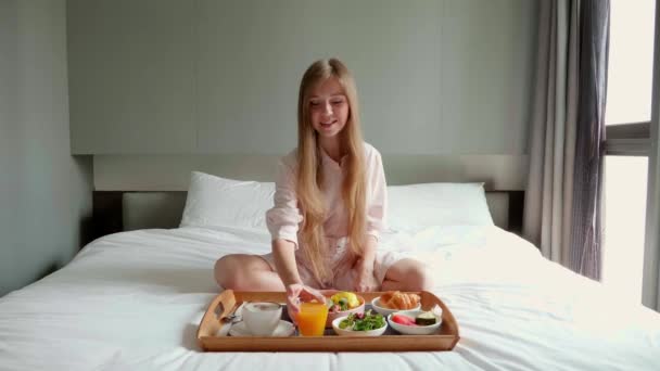 Joven sonriente hermosa mujer comiendo desayuno en la cama en acogedora habitación de hotel — Vídeos de Stock