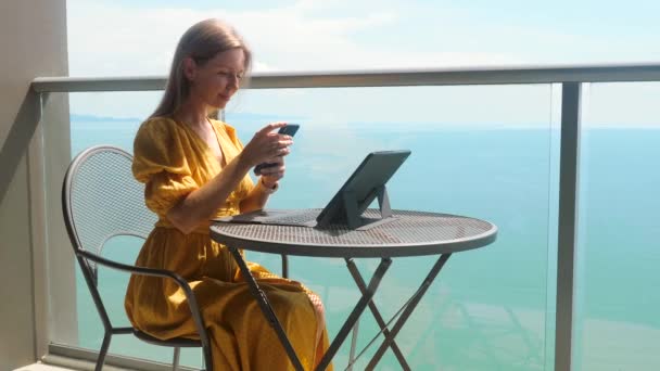 Zakelijke vrouw met mobiele telefoon en tablet zitten aan tafel met zeezicht — Stockvideo