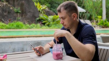 Modern Genç Adam Akıllı Telefon Kullanıyor, E-postaları Kontrol Ediyor, Çevrimiçi Haberlere Bakıyor, Pembe Sağlıklı Meyve Smoothie İçiyor, Vegan 'la Tropikal Kafe' de Oturuyor, Havuz kenarındaki Vejetaryen Yemeği Yiyiyor. Detox Uygun Beslenme, Konsept.