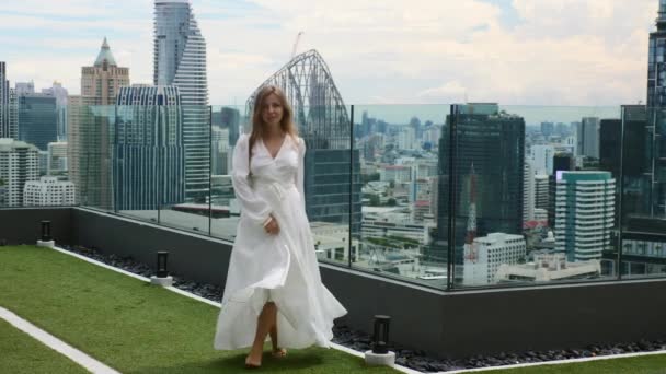 Χαμογελαστή γυναίκα με κομψό λευκό φόρεμα μόδας Περπατώντας στην οροφή με ουρανοξύστες — Αρχείο Βίντεο
