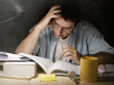 Young student at home desk reading biting pen studying at night with pile of books and coffee clipart