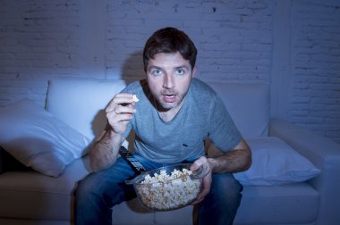 young attractive man at home lying on couch watching tv holding popcorn bowl eating