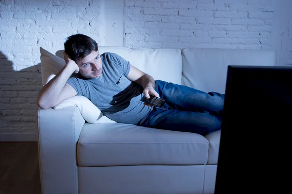 Young attractive man at home lying on couch at living room watching tv holding remote control — Zdjęcie stockowe