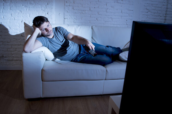 young attractive man at home lying on couch at living room watching tv holding remote control