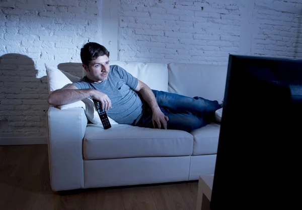 Jovem atraente homem em casa deitado no sofá na sala de estar assistindo tv segurando controle remoto — Fotografia de Stock
