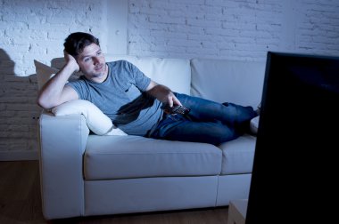 young attractive man at home lying on couch at living room watching tv holding remote control