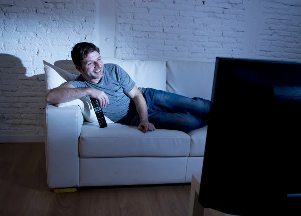 Jovem atraente homem em casa deitado no sofá na sala de estar assistindo tv segurando controle remoto — Fotografia de Stock