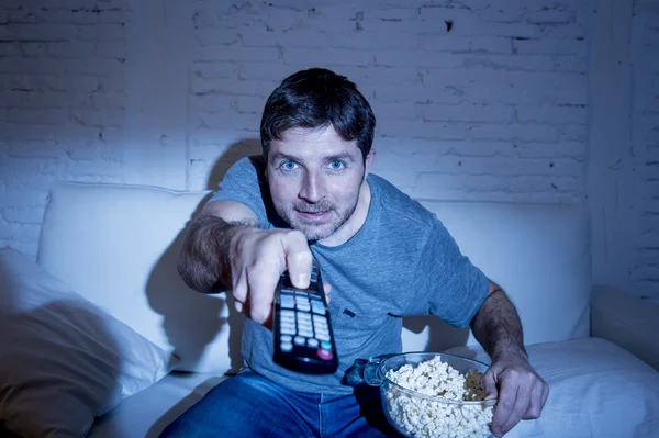 Hombre en casa acostado en el sofá en la sala de estar viendo la televisión comer palomitas de maíz bowl usando control remoto —  Fotos de Stock