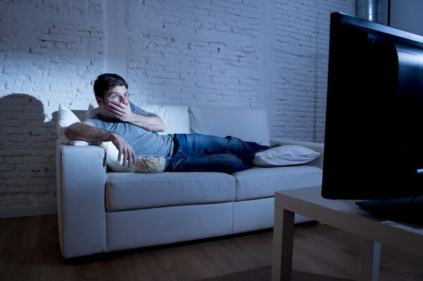 attractive man at home lying on couch at living room watching tv  eating popcorn bowl looking surprised