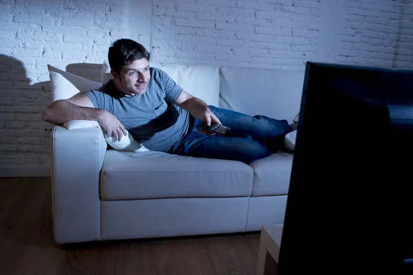 Молодой привлекательный счастливый человек лежит на диване в гостиной смотреть возбужденный телевизор с пультом дистанционного управления — стоковое фото