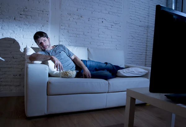 Attrayant homme à la maison couché sur le canapé au salon regarder la télévision manger bol de maïs soufflé regardant surpris — Photo