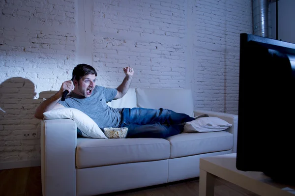 Attractive man at home lying on couch at living room watching sport match on tv celebrating goal — Stock Photo, Image