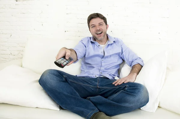 Молодой счастливый человек смотрит телевизор улыбаясь и смеясь на диване — стоковое фото