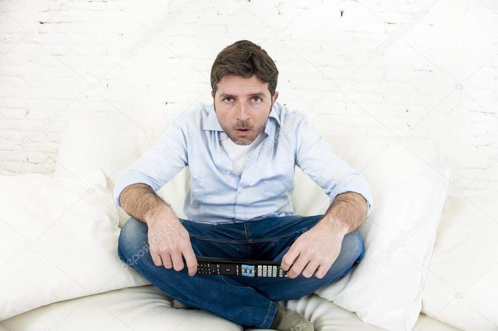 man watching television at home living room sofa with remote control looking very interested