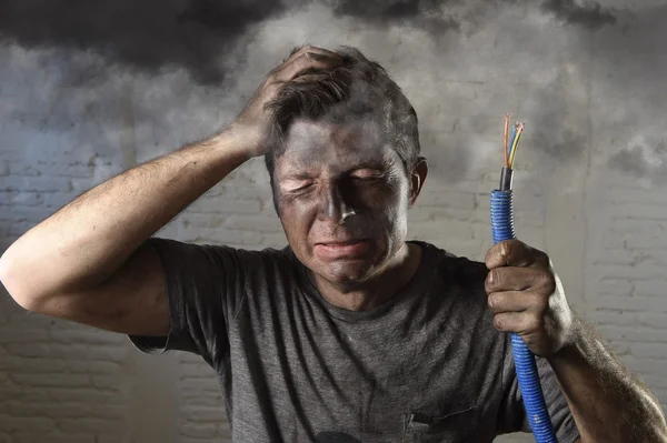 Młody człowiek trzymając kablówkę palenia po wypadku elektrycznym z brudną spaloną twarzą w zabawny smutny wyraz twarzy — Zdjęcie stockowe