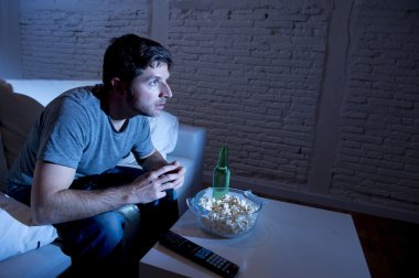 young television addict man sitting on home sofa watching TV eating popcorn and drinking beer bottle