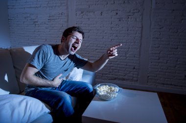 young television addict man sitting on home sofa watching TV and eating popcorn laughing crazy