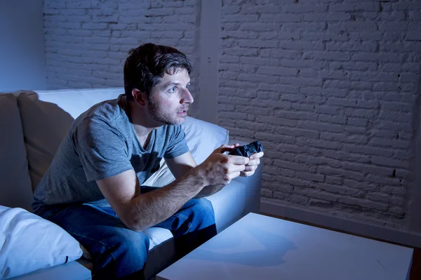 Young excited man at home sitting on living room sofa playing video games using remote control joystick — Stockfoto