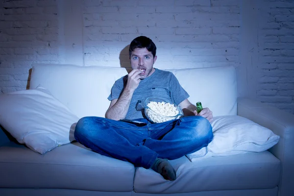 Young television addict man sitting on home sofa watching TV eating popcorn and drinking beer bottle — Stockfoto