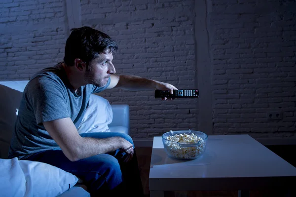 Adicto a la televisión hombre sentado en el sofá de casa viendo la televisión comer palomitas de maíz usando control remoto —  Fotos de Stock